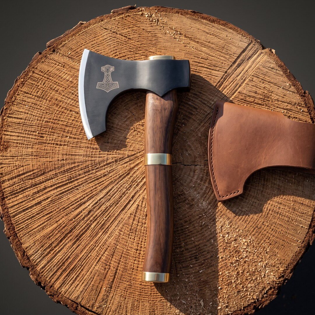 Mjolnir bearded axe with a blackened 1095 steel head and brass banded rosewood haft resting on a cut log round beside it Mjolnir bearded axe with a blackened 1095 steel head and brass banded rosewood haft resting on a cut log round beside its brown leather blade cover.