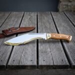 Mjolnir bolster kukri knife in polished 1095 steel with figured olive wood scales and brass fittings resting on weathered grey decking beside its brown leather sheath