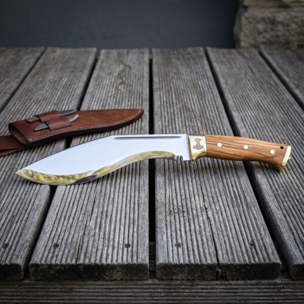 Mjolnir bolster kukri knife in polished 1095 steel with figured olive wood scales and brass fittings resting on weathered grey decking beside its brown leather sheath