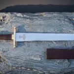Mjolnir etched arming sword in bright polished high carbon steel with a brass crossguard, oxblood leather grip, brass brazil nut pommel and leather scabbard on slate rock