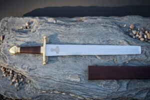 Mjolnir etched arming sword in bright polished high carbon steel with a brass crossguard, oxblood leather grip, brass brazil nut pommel and leather scabbard on slate rock