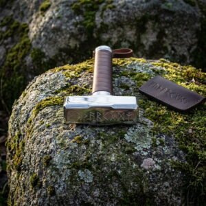 Mjolnir war hammer with a polished steel head cut with Elder Futhark runes and a tan leather wrapped haft, resting on a mossy boulder beside its leather cover
