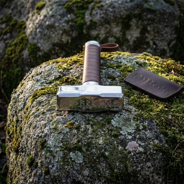 Mjolnir war hammer with a polished steel head cut with Elder Futhark runes and a tan leather wrapped haft, resting on a mossy boulder beside its leather cover