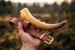 Natural ox drinking horn with polished brass rim held in one hand with a tan leather belt hanger stamped with a Vegvisir