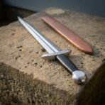 Norse dagger with smooth black grip lying diagonally across a sandstone block, tan leather sheath running alongside
