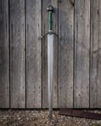 Norse damascus sword with a green wrapped grip, blackened scrolled crossguard and Mjolnir pommel standing point down on gravel against weathered wooden planks