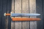Norse sword with brass guard and pommel and tan leather grip laid on dark boards above its full length tan leather scabbard