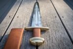 Norse sword with curved brass guard, brazil nut brass pommel and tan leather grip on wooden decking beside its leather scabbard