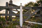 Norse sword with rune engraved blade, brass cross and bone white grip standing point down by a wooden field gate