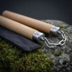 Oak nunchaku batons resting side by side on a lichen covered rock, steel chain looped between the engraved caps, pouch alongside