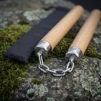 Close up of oak nunchaku end caps with triskele engraving and polished steel chain on mossy rock, black pouch behind