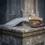 Olive wood damascus kukri with a brass bolster and etched vegvisir resting on a weathered stone plinth beside its stitched brown leather sheath and companion blades.