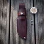 Overhead view of an oxblood leather knife sheath with raven tooling and snap strap on grey decking with balm tin and studs