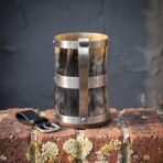 Ox horn mug with brushed steel rune band and tall steel handle standing on an old brick wall beside a black leather hanger