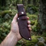 Oxblood leather knife sheath with raven tooled face and hand stitched welt held upright in a hand in a mossy forest