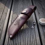 Oxblood leather knife sheath with tooled raven panel and snap strap on decking beside spare snap studs and a tin of leather balm