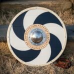 Painted wooden round shield with cream rawhide rim and navy pinwheel standing upright in grass beside a leather strap