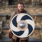 Painted wooden round shield with navy pinwheel and steel boss held at waist height by a bearded man on stone steps
