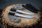 Pair of hand forged chain knives laid on a tree stump, full length side view with steel chains and two black sheaths