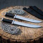 Pair of hand forged chain knives laid on a tree stump, full length side view with steel chains and two black sheaths