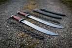Pair of hand forged chain swords with oxblood grips set diagonally on a gravel path, blackened chains and two scabbards