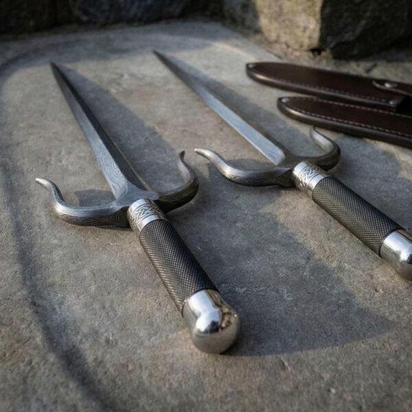 Pair of hand forged Damascus sai daggers, low close view of the side prongs and steel pommels with sheaths behind