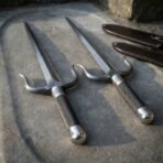 Pair of hand forged Damascus sai daggers laid parallel on a carved stone slab, two leather sheaths in the top corner