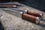 Pair of hand forged seax knives with leather grips and steel bolsters lying on a stone ledge, two sheaths behind