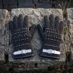 Pair of blackened butted chainmail gauntlets with domed steel studs and white leather buckle straps laid on a weathered stone block outdoors