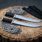 Pair of hand forged 1095 steel rune knife blades etched with Elder Futhark on ring pommel chains resting on a cut tree stump