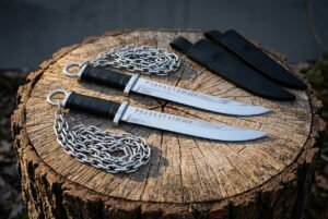 Pair of hand forged 1095 steel rune knife blades etched with Elder Futhark on ring pommel chains resting on a cut tree stump