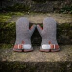 Pair of hand forged chainmail mittens in bright mild steel butted mail with russet leather cuffs and engraved knotwork buckle plates resting on a mossy stone wall