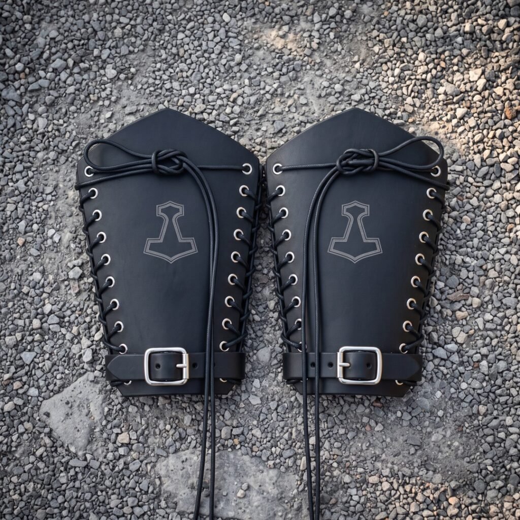 Pair of hand made black leather bracers with engraved Mjolnir hammers, steel eyelet lacing and buckled wrist straps, laid out on grey gravel