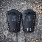 Pair of hand made black leather bracers with engraved Mjolnir hammers, steel eyelet lacing and buckled wrist straps, laid out on grey gravel