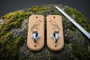 Pair of hand made oak sword wall mount plaques engraved with standing ravens, each fitted with a brushed steel hook, resting on mossy stone beside a polished blade.