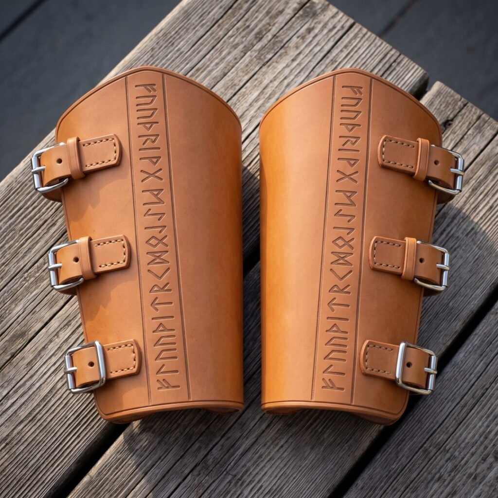 Pair of hand made tan leather bracers embossed with an Elder Futhark rune line and closed by three steel roller buckles, resting on weathered grey timber decking.