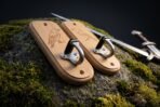 Pair of oak sword wall hooks with engraved ravens and brushed steel hooks resting on a mossy rock beside a sword