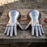 Pair of polished steel plate gauntlets with articulated finger lames and knotwork etched bell cuffs on a weathered wooden table beside brown leather gloves