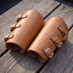Pair of tan leather bracers embossed with a rune line and fitted with three steel roller buckles each, on a wooden table
