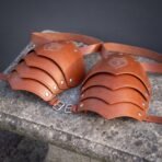 Pair of tan leather pauldrons with riveted articulated lames, buckled straps and wolf head engraving on a stone bench