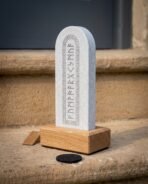 Pale grey slate rune stone with arched knotwork border and elder futhark row standing in a solid oak base on a stone step
