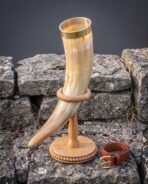 Pale natural ox horn drinking horn with brass rim band and etched valknut on a beaded oak stand, tan leather belt beside it