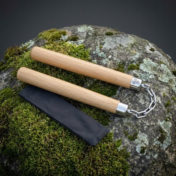 Pale oak hardwood nunchaku with polished steel end caps, a triskele engraving and a heavy steel chain lying on a mossy lichen covered boulder beside a black pouch