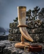 Pale ox horn drinking horn with brass rim on an oak ring stand set on a misty stone wall, tan leather belt with brass buckle