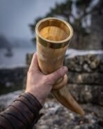 Pale ox horn drinking horn with solid brass rim band and etched valknut held up in one hand, misty trees and water behind