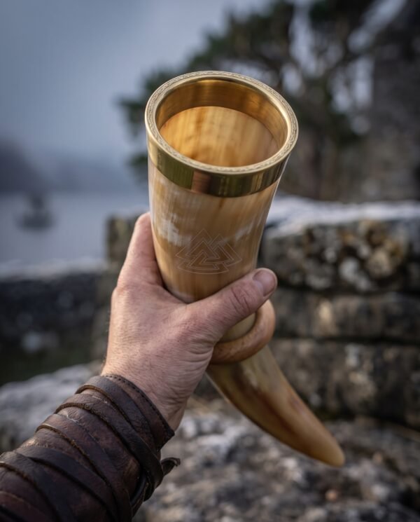 Pale ox horn drinking horn with solid brass rim band and etched valknut held up in one hand, misty trees and water behind