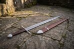 Pattern welded arming sword with brown leather grip and steel wheel pommel lying on flagstones beside its leather scabbard