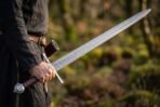 Pattern welded arming sword with etched knotwork fuller held at the side by a man in a dark linen tunic in the woods
