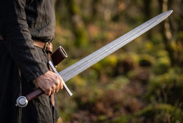 Pattern welded arming sword with etched knotwork fuller held at the side by a man in a dark linen tunic in the woods