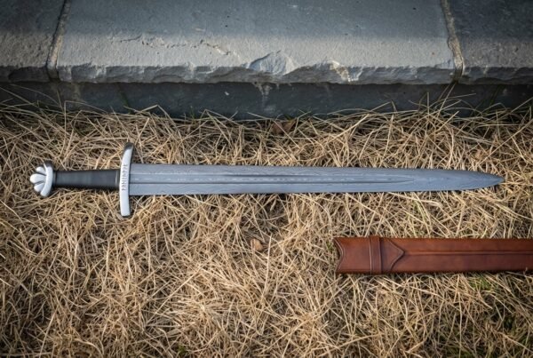 Pattern welded Damascus five lobed viking sword with a black leather grip and runes etched at the ricasso lying on dry grass beside a stone kerb and leather scabbard
